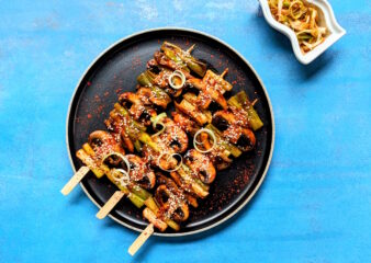 Gochugaru Pressed Mushroom Dakkochi Skewers on a dark plate against a bright blue background, prepared in the Kick Start Korean class at COOK! with the Vegetarian Society.